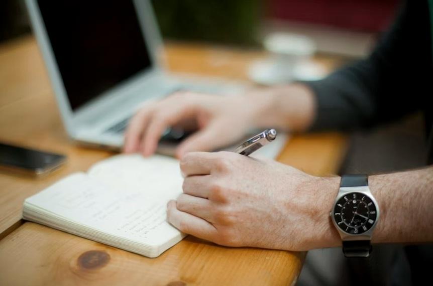 Person writing a list next to a laptop.